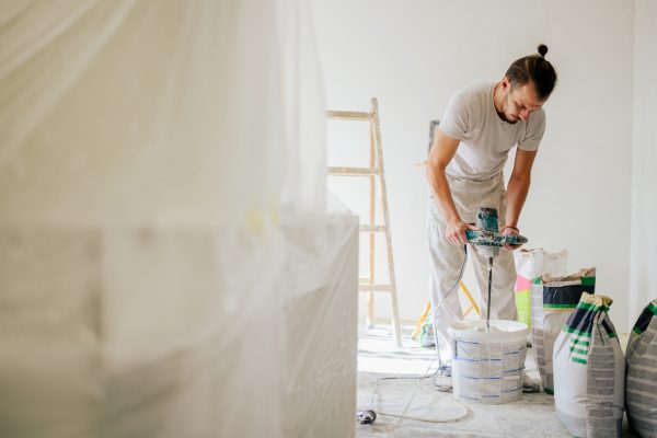 A constructor is standing in a house in a renovation process and mixing plaster for renovating and skim coating walls. Full length of a worker mixing materials for plastering walls. A plasterer works.
